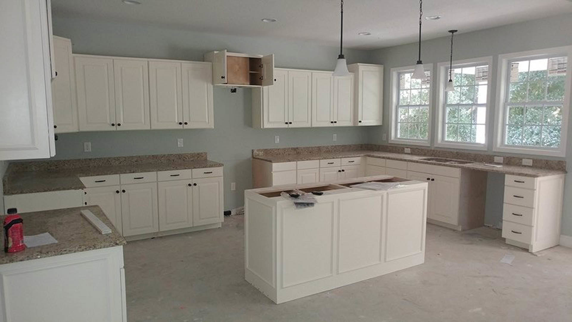 White kitchen with shaker cabinets, central island with drawers, stainless steel appliances, row of windows with white trim, person in red jacket blurred in background