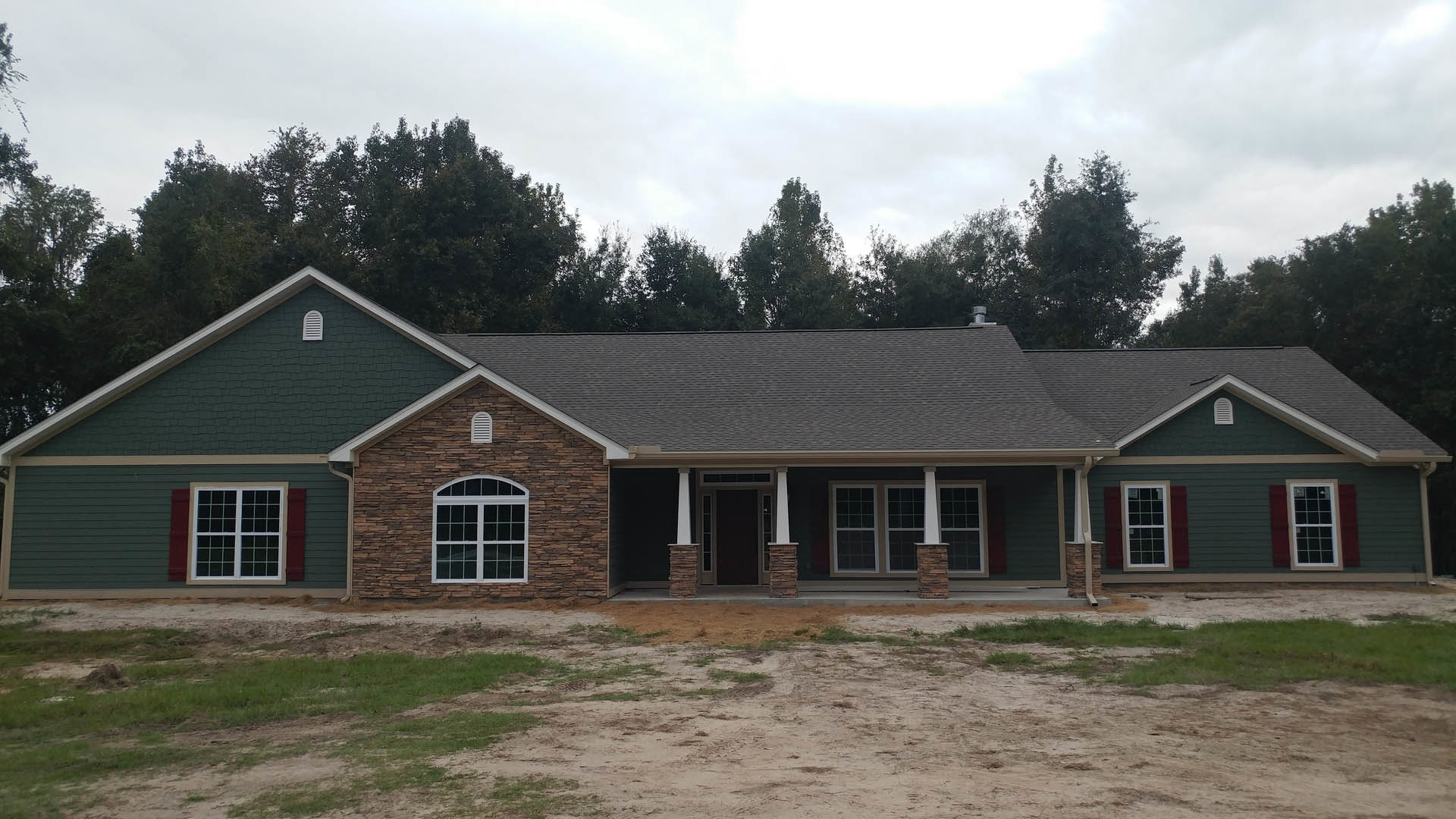 Two-story house with stone and siding exterior, multiple white-framed windows, dark roof, and expansive grassy yard bordered by trees under a partly cloudy sky