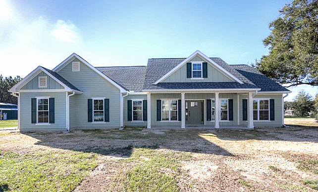 Spacious front porch with white railings, multiple windows with white frames, light-colored siding, manicured lawn, white pole, leafy tree, and clear blue sky