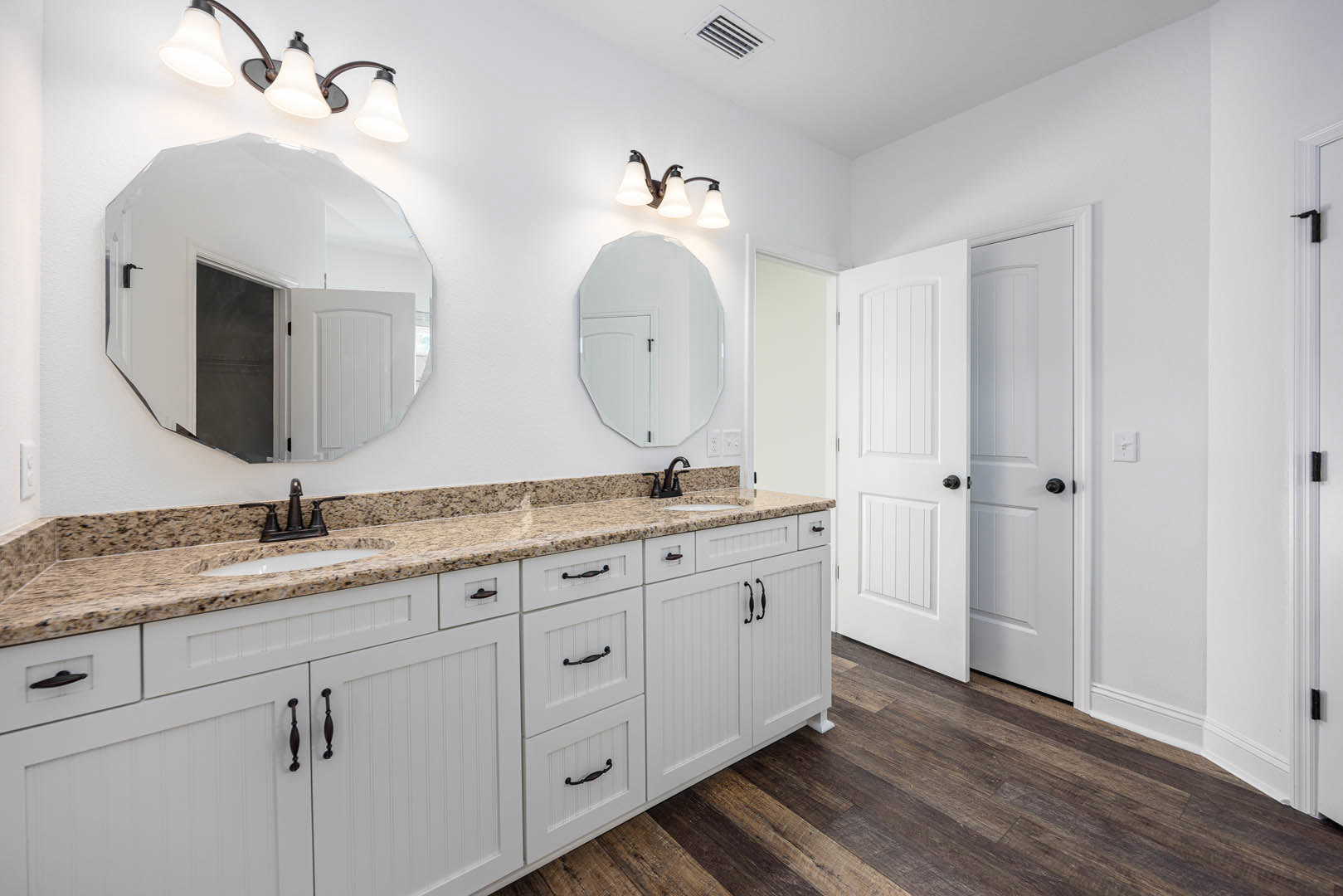White bathroom cabinets with marble countertops, rectangular wall mirror above sink, black-handled white door, three-light fixture mounted above mirror, chrome faucet, tile