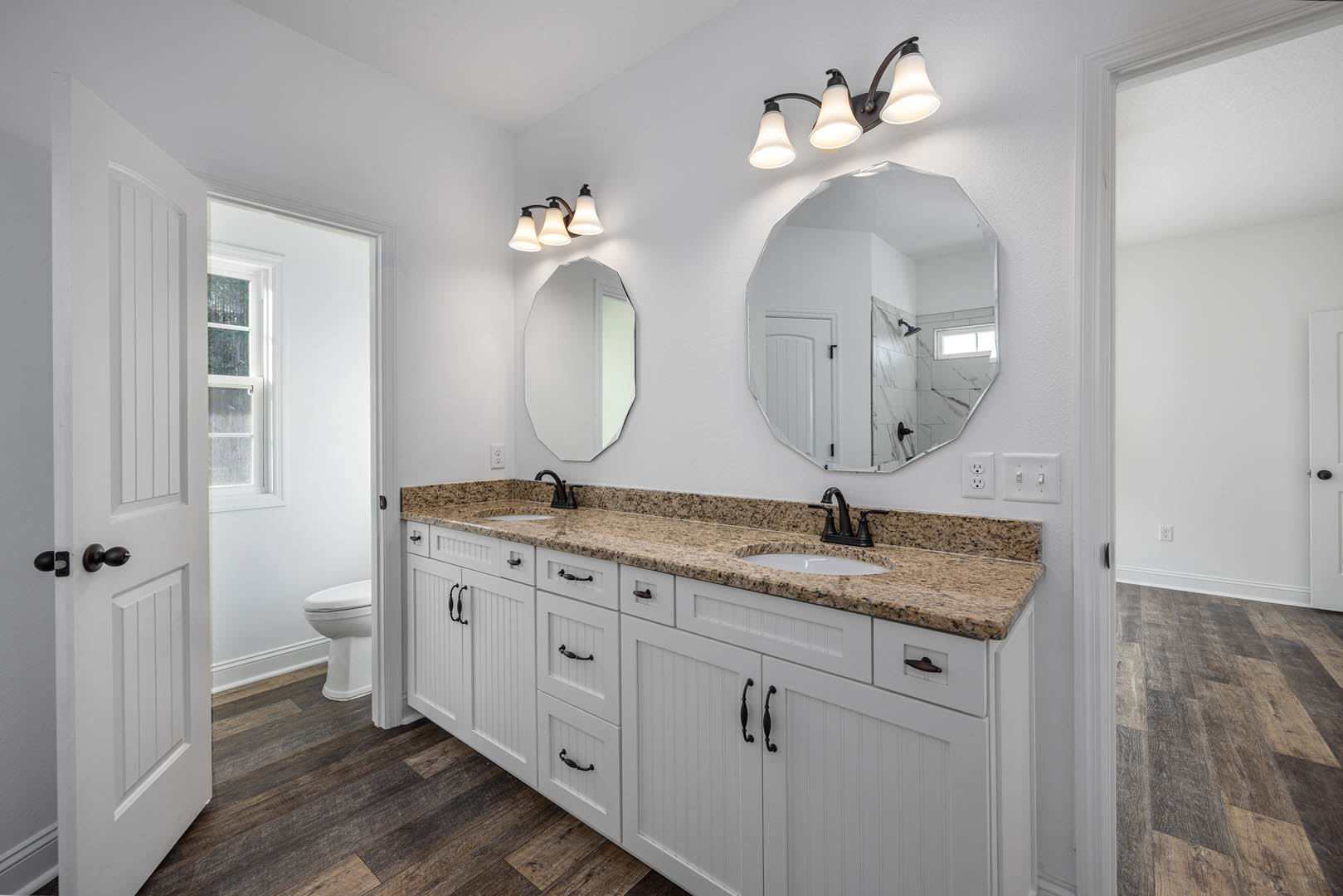 Bathroom with white shaker cabinets, marble countertops, rectangular mirror with black frame, three-light fixture above, white toilet, and wood flooring