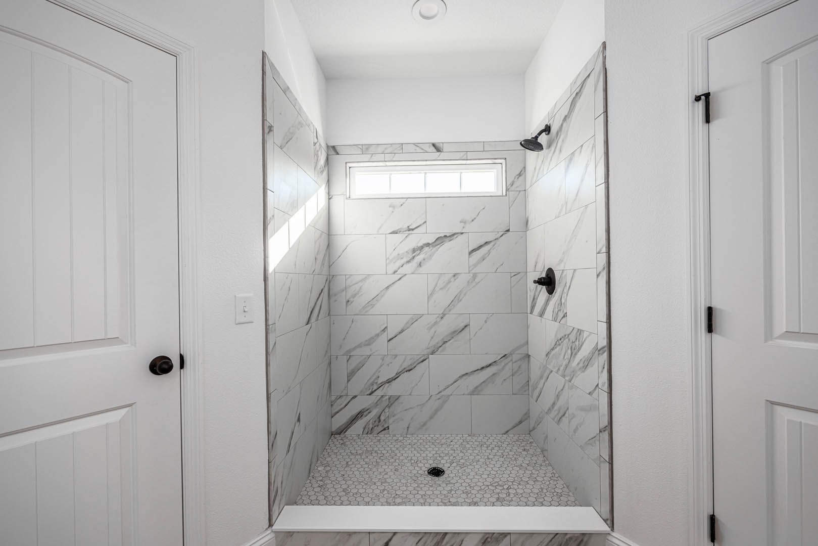 Marble tile shower with white tile floor and linear drain, white door with silver knob, window, and contrasting black grout lines