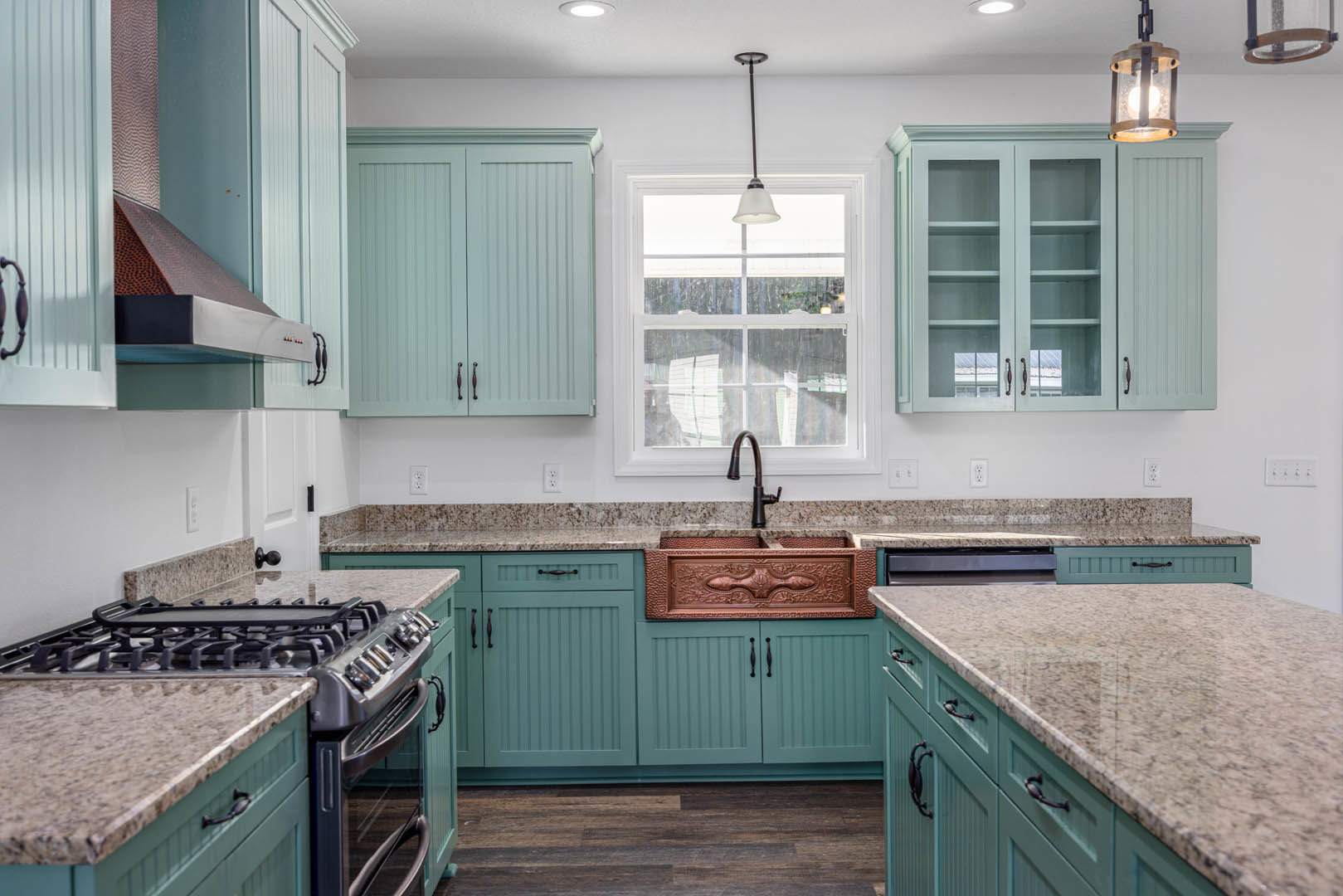 Blue kitchen cabinets with glass doors, granite countertops, stainless steel sink and stovetop, white window, tile backsplash, and modern light fixture.