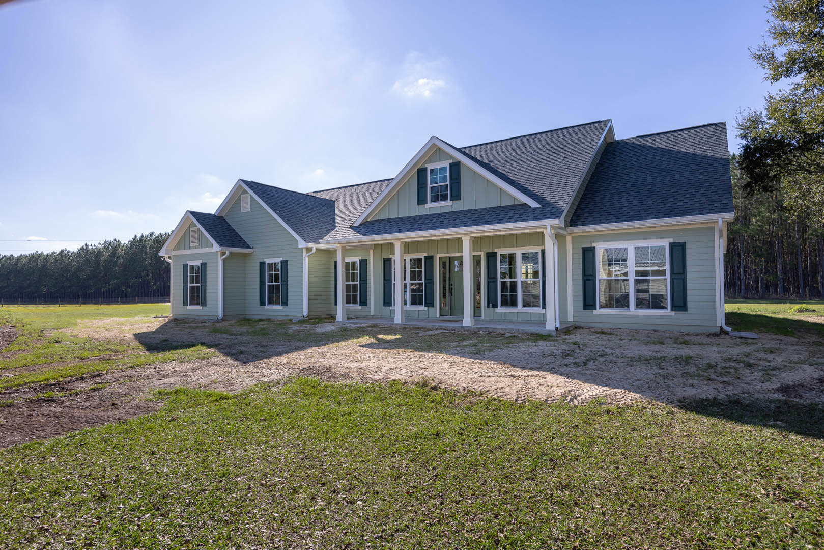 Two-story home with green roof, white-framed windows, covered porch, and manicured front lawn bordered by mature trees under partly cloudy sky