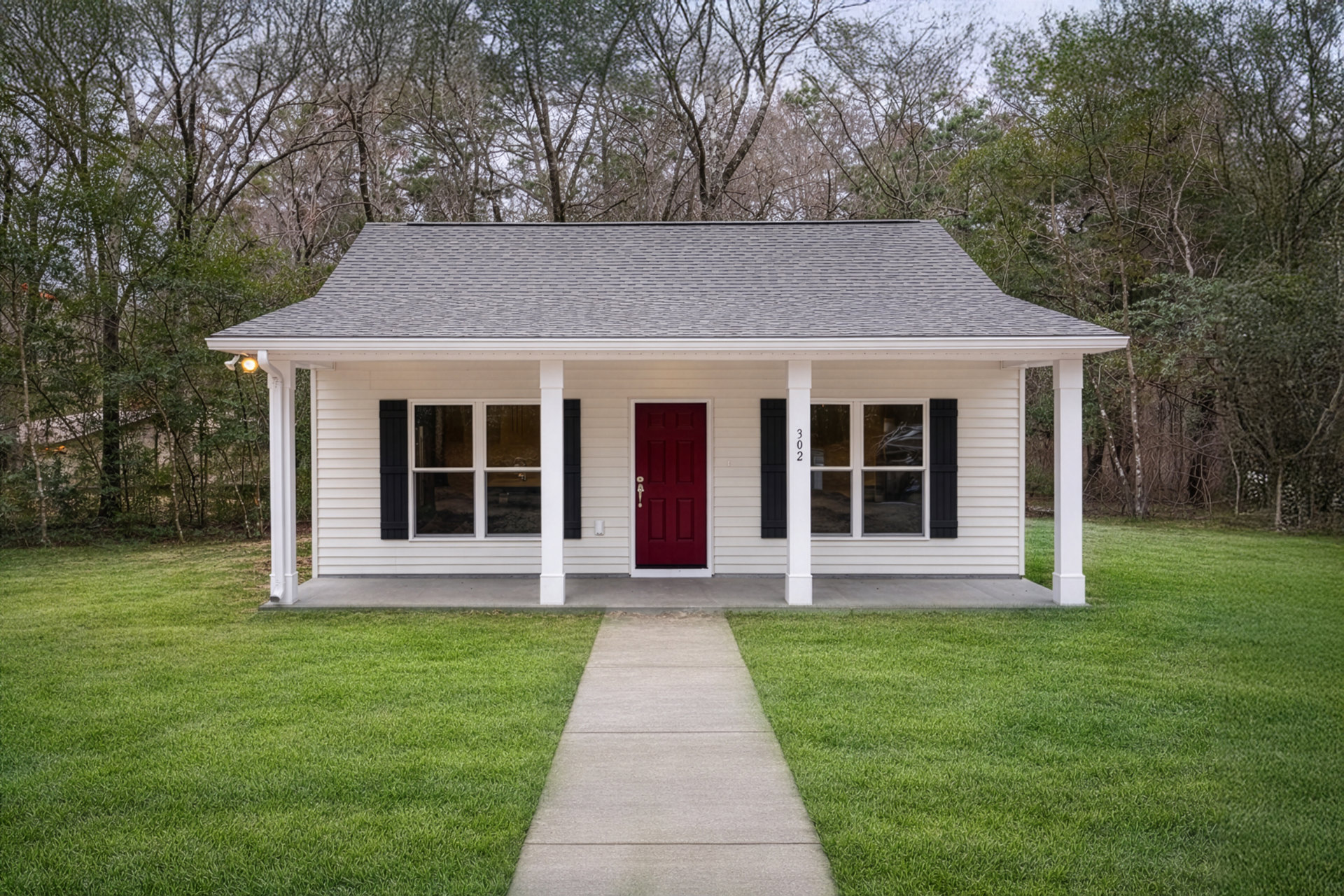 White siding house with black shutters, red front door framed in white, concrete walkway bordered by green grass, mature tree and shrubs in front yard.
