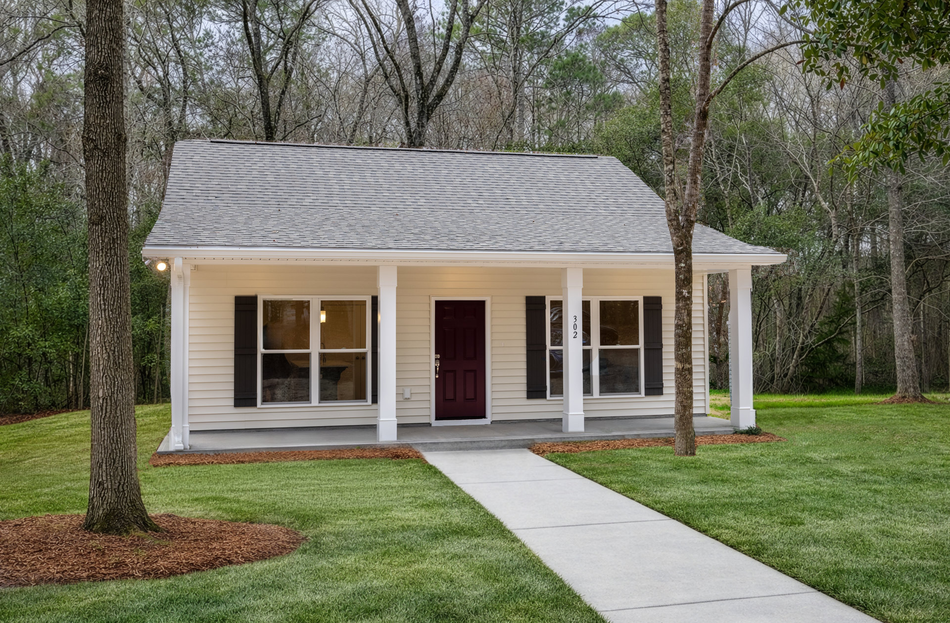 White siding house with red front door, white columns on porch, lit window, concrete walkway, tree trunk with brown mulch, green grass in yard
