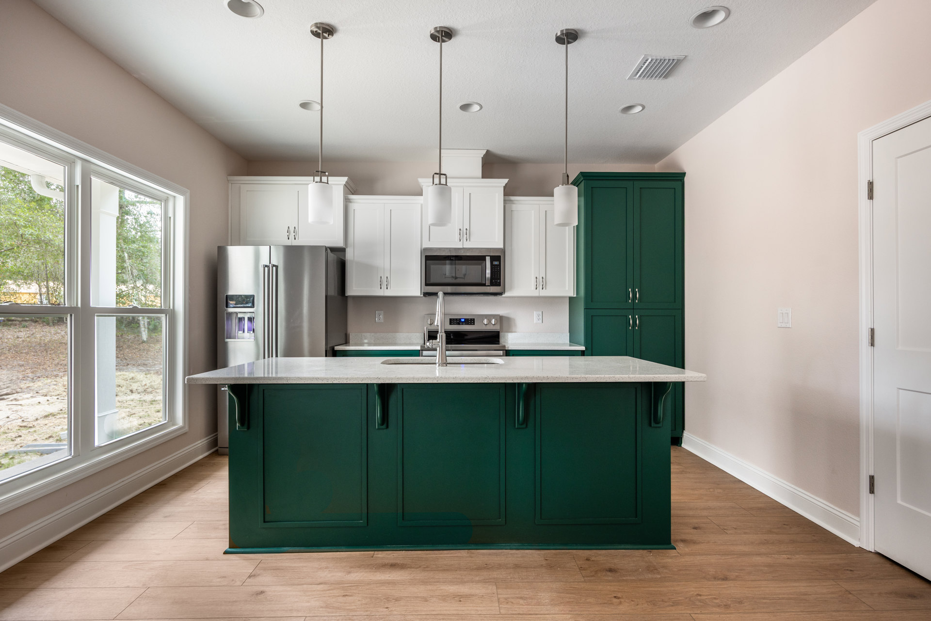 Green lower cabinets and white upper cabinets in a kitchen with a silver double-door refrigerator, built-in microwave, white door, stainless steel sink and faucet, window