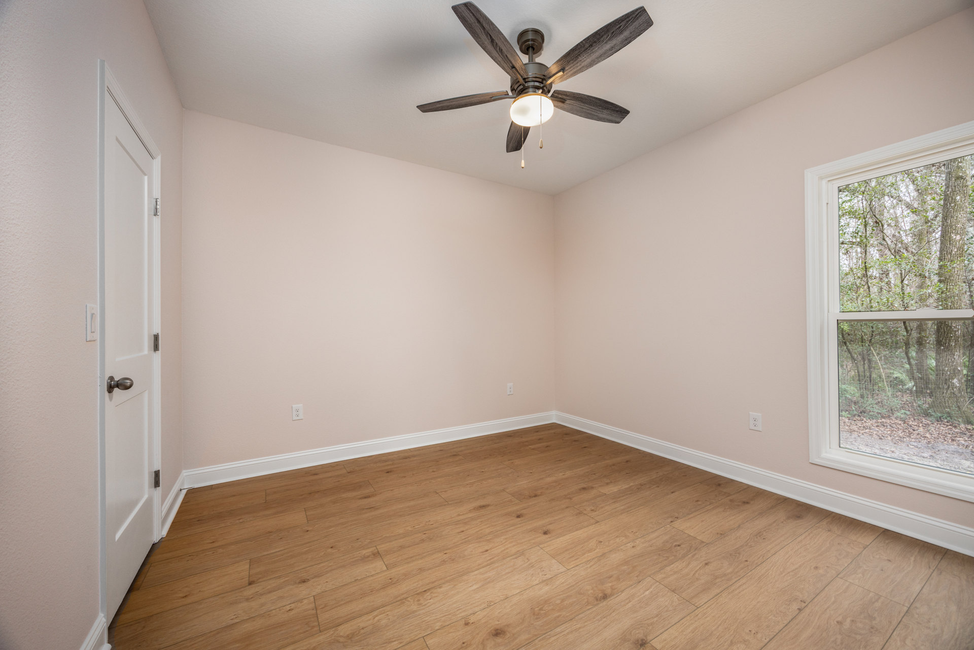 Ceiling fan with integrated light fixture mounted on white plaster ceiling, wood laminate flooring, large window revealing trees and dirt outside