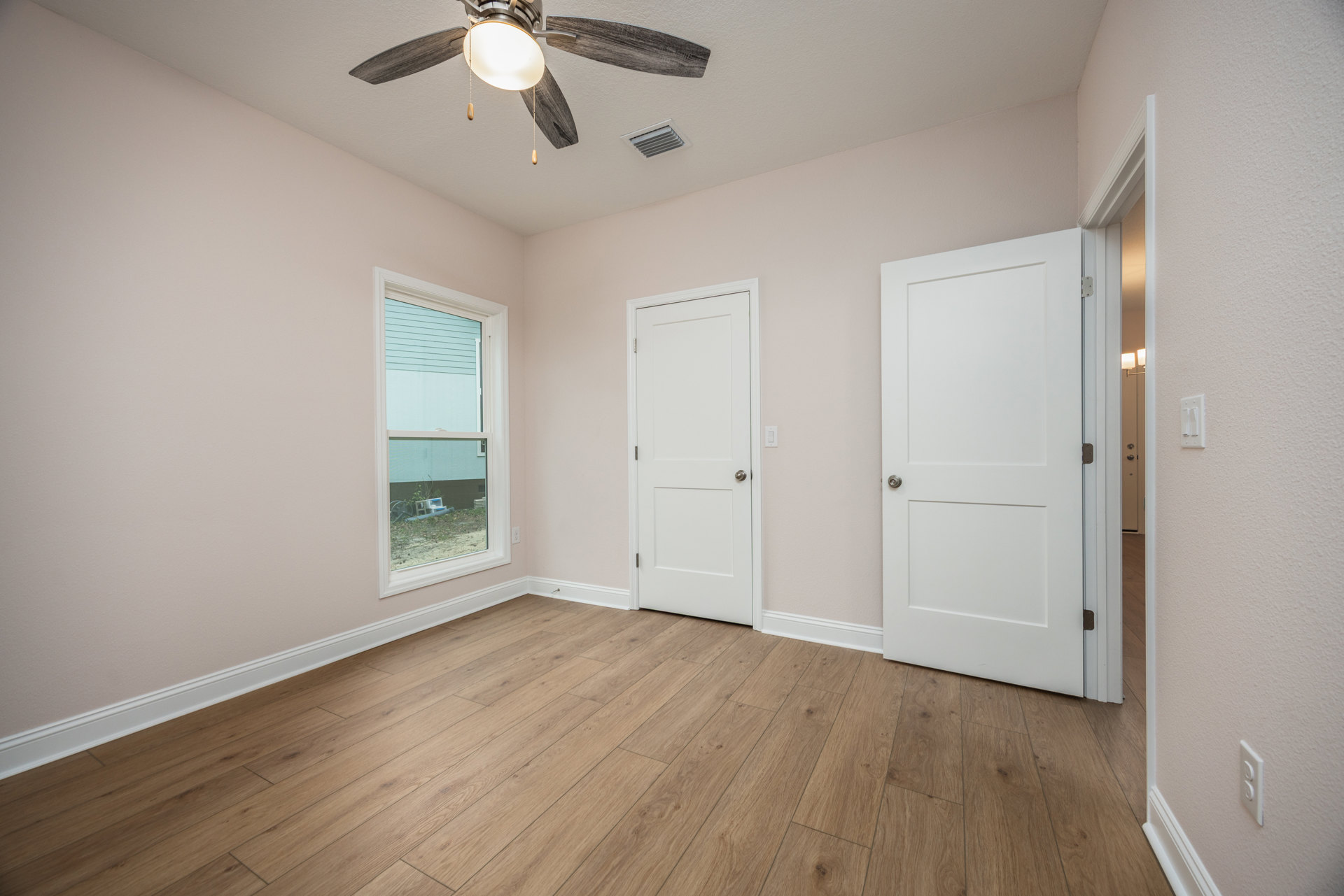 Wood floor room with white walls, two white doors with silver knobs, window with white trim, ceiling fan with light fixture.