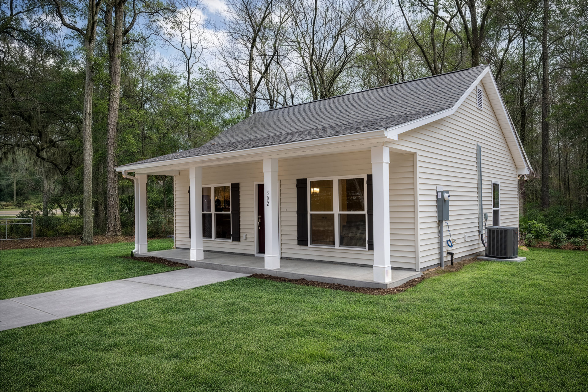 Two-story house with white siding, covered front porch, large windows, black heat pump unit, concrete walkway, green lawn, and mature trees in the background