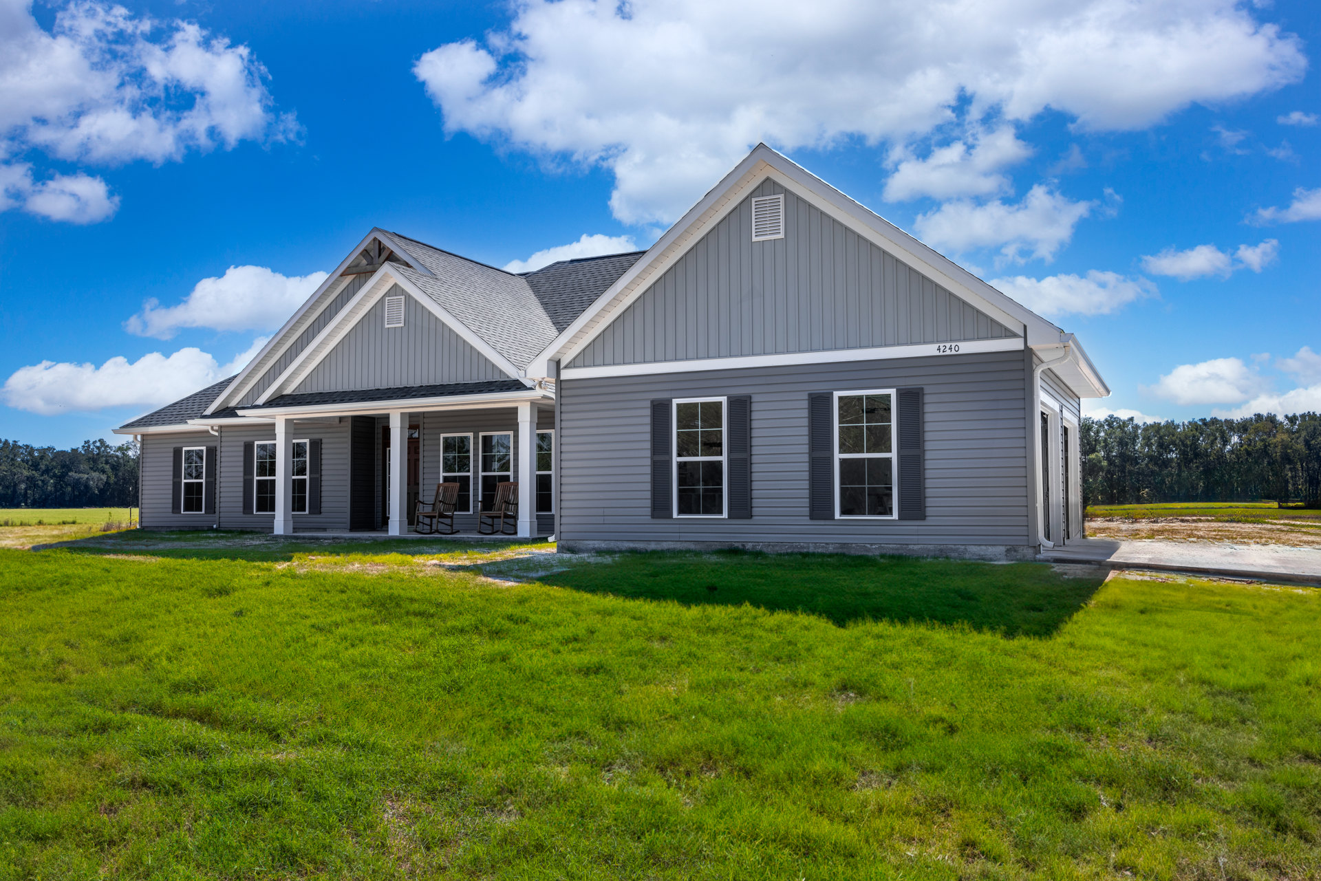 Two-story house with white-framed windows, gray shingle roof, green grass lawn, outdoor chairs, and blue sky with scattered clouds