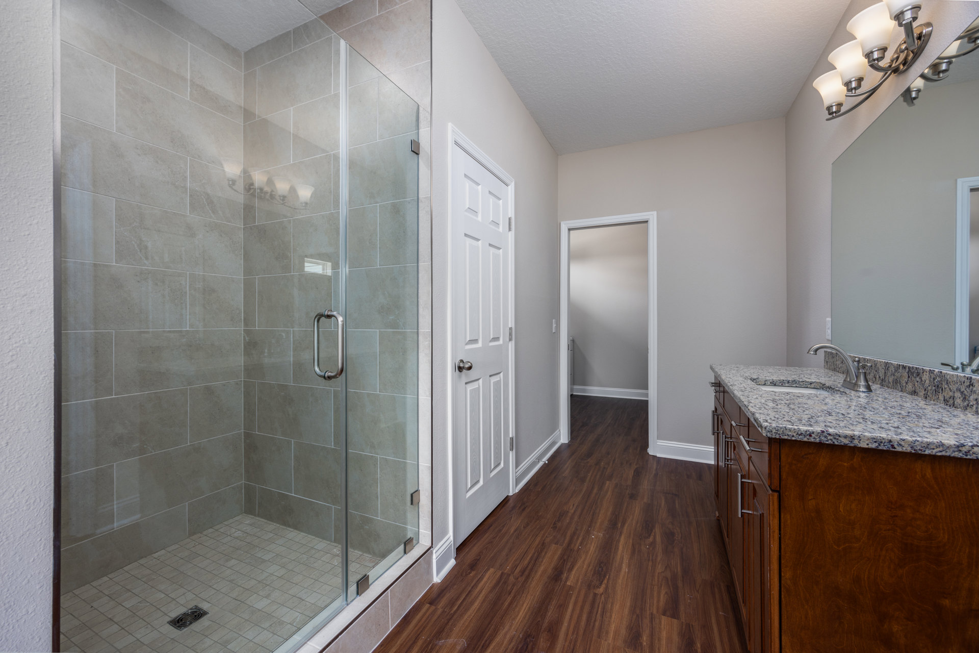 Bathroom with glass shower enclosure, wood flooring, wall-mounted sink and mirror, metal drain on tile, white-shaded light fixture, and chrome shower handle.