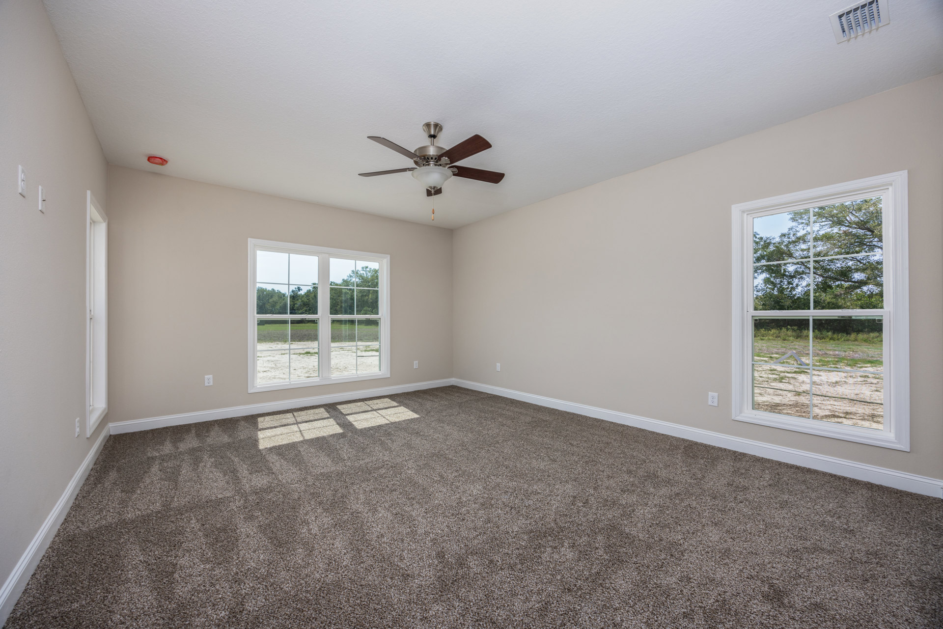 Carpeted room with white walls, ceiling fan with light fixture, large window overlooking trees, wall vent visible