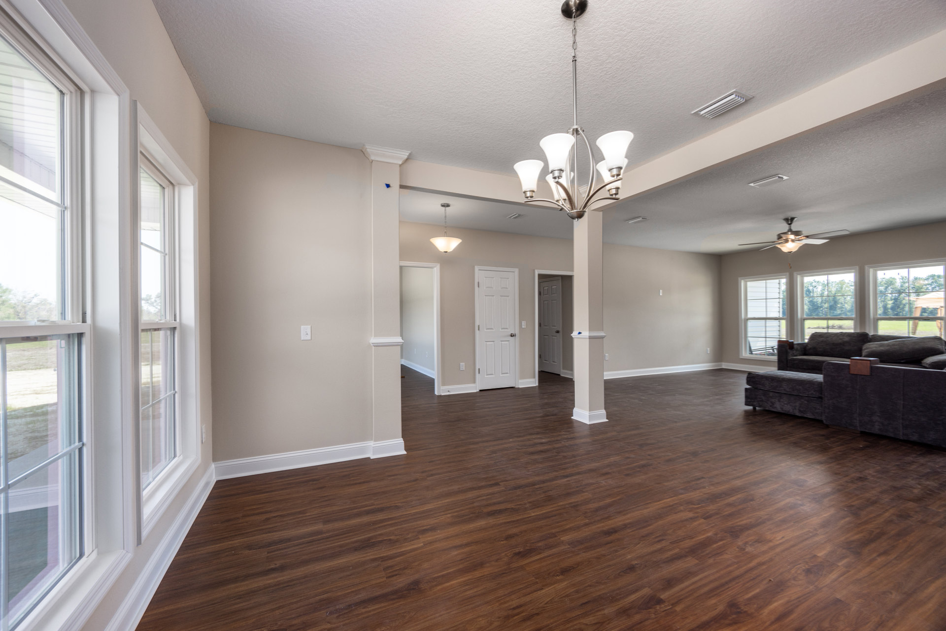 Spacious living area with hardwood floors, white columns, ceiling fan, white-framed window, white door with silver knobs, and a couch