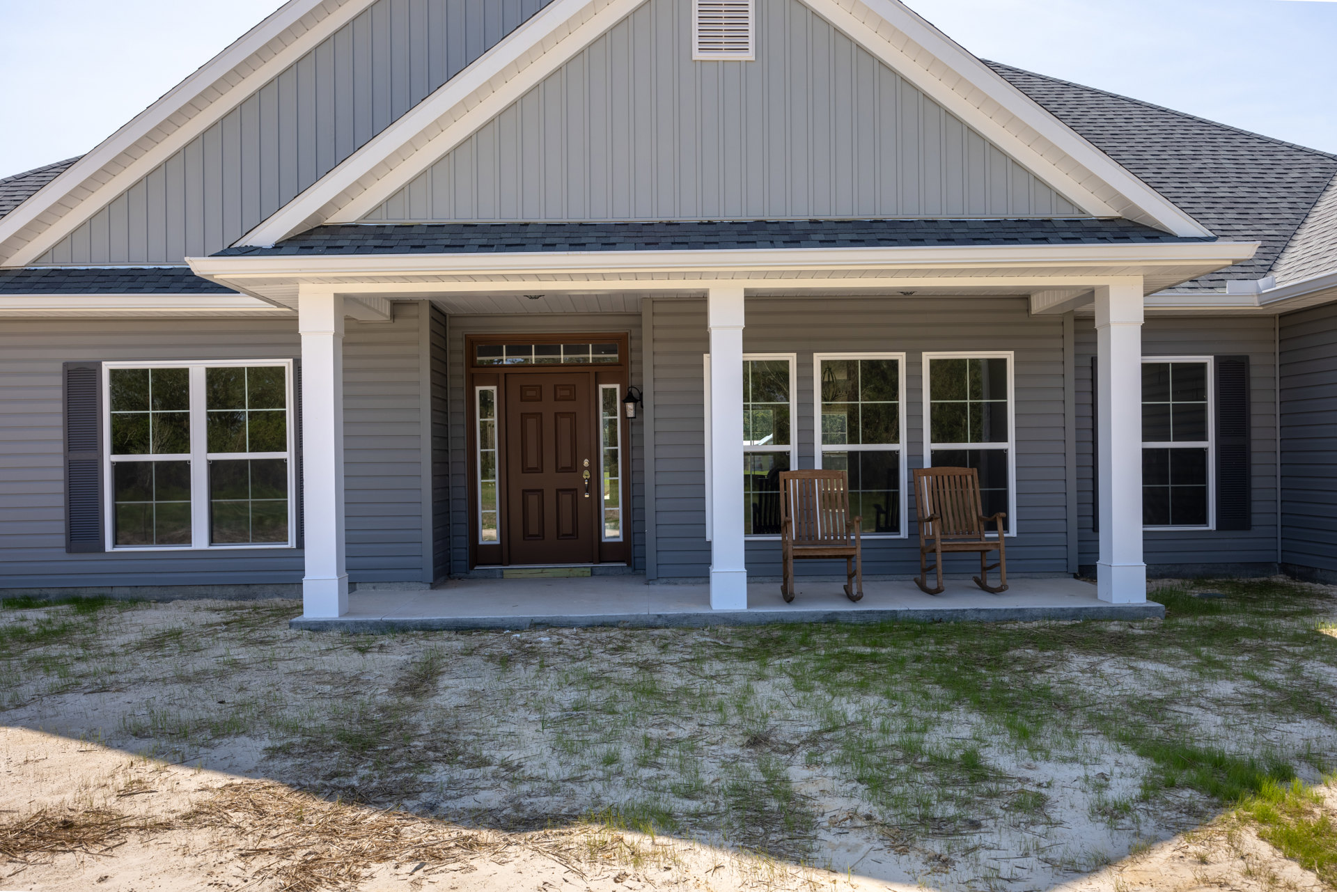 Front porch with two wooden chairs, brown door with glass panes, white-framed window, light-colored siding, patch of grass and dirt in foreground