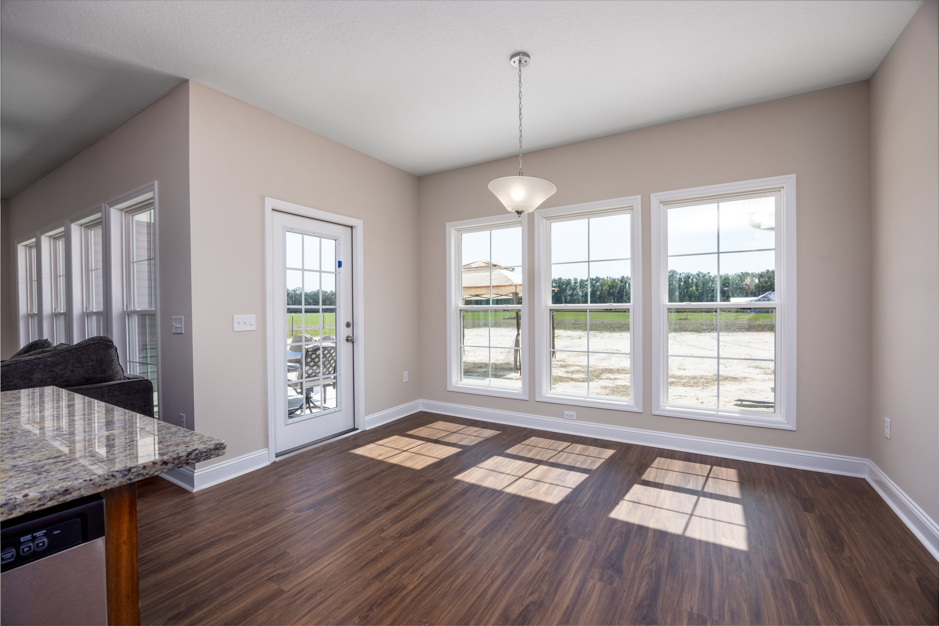Open living area with wood flooring, white-trimmed windows, marble coffee table in front of a couch, kitchen countertop with built-in dishwasher, ceiling chain light fixture, and
