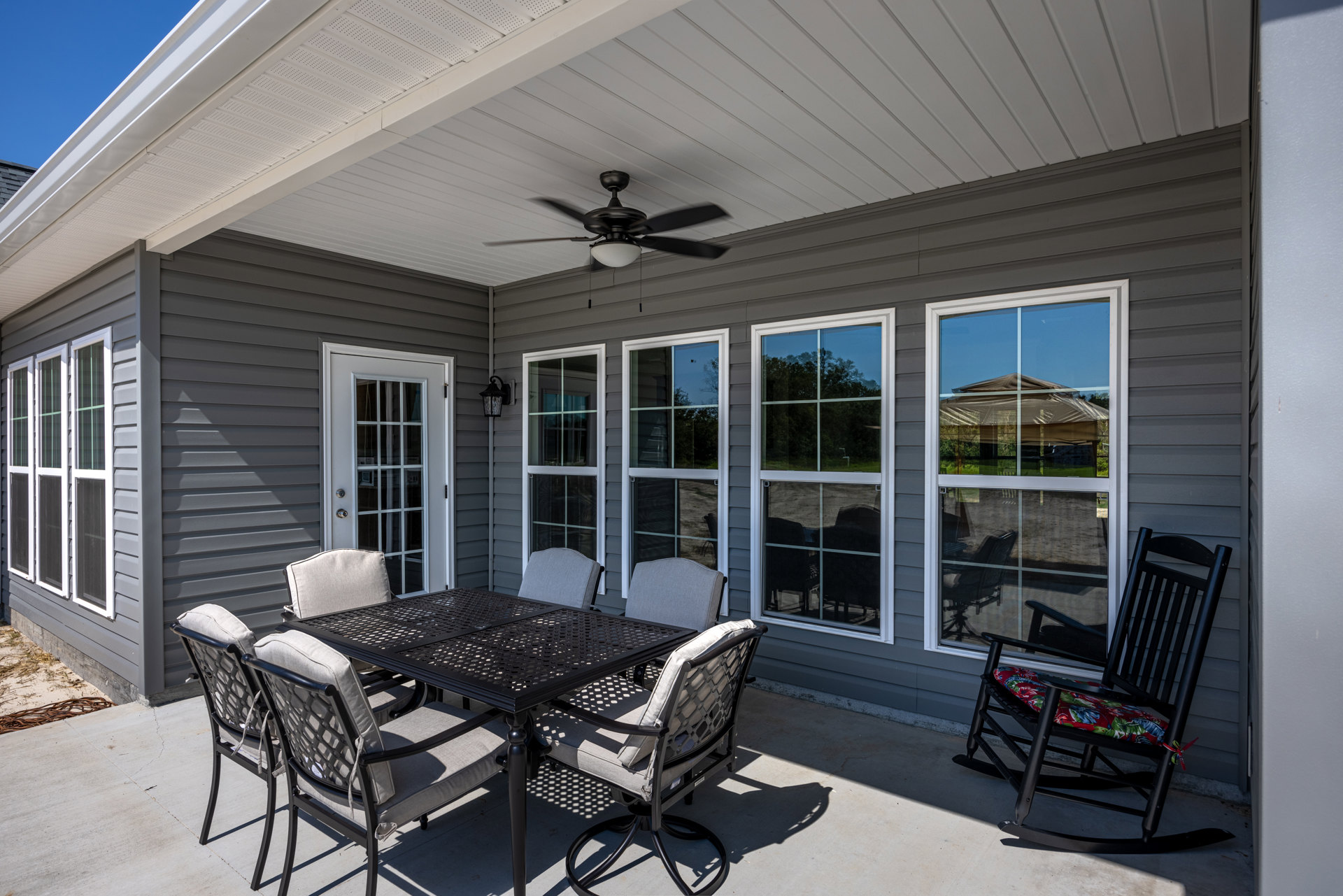 Patio with metal table and four chairs, rocking chair beside window, glass-paneled door, ceiling fan overhead, light-colored siding and deck flooring