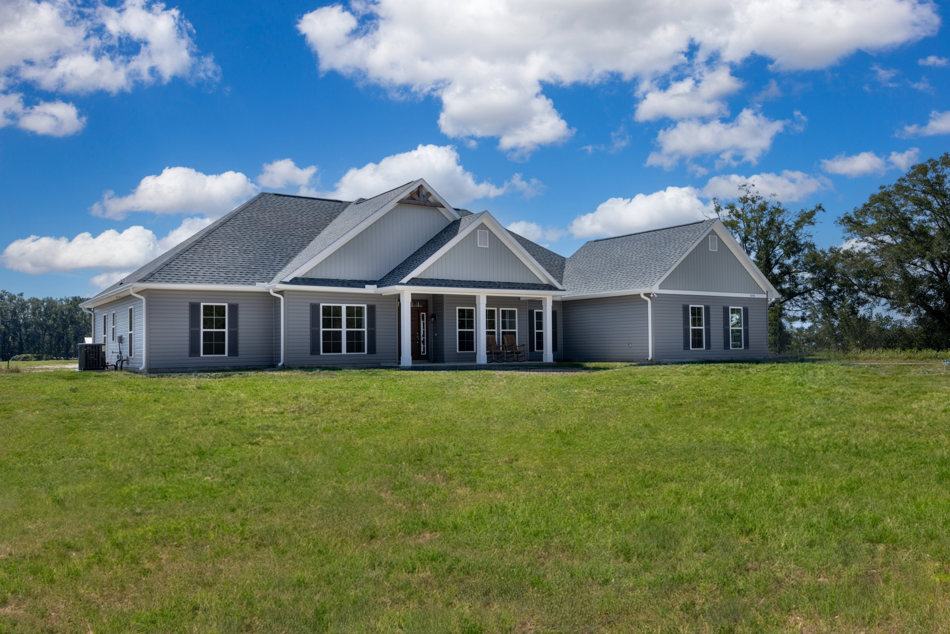 Two-story house with gray siding, covered front porch featuring white rocking chairs, manicured green lawn, mature tree, blue sky with scattered clouds, air conditioning unit