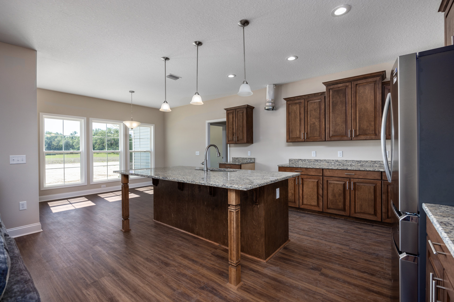 Spacious kitchen featuring a large central island with built-in sink, white countertops, modern cabinetry, stainless steel appliances, and contemporary ceiling light fixtures.