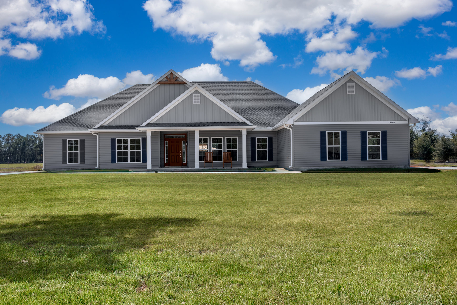 White farmhouse-style home with gray roof, brown glass-paneled front door, covered porch featuring two wooden chairs, expansive green lawn, large windows, and partly cloudy sky