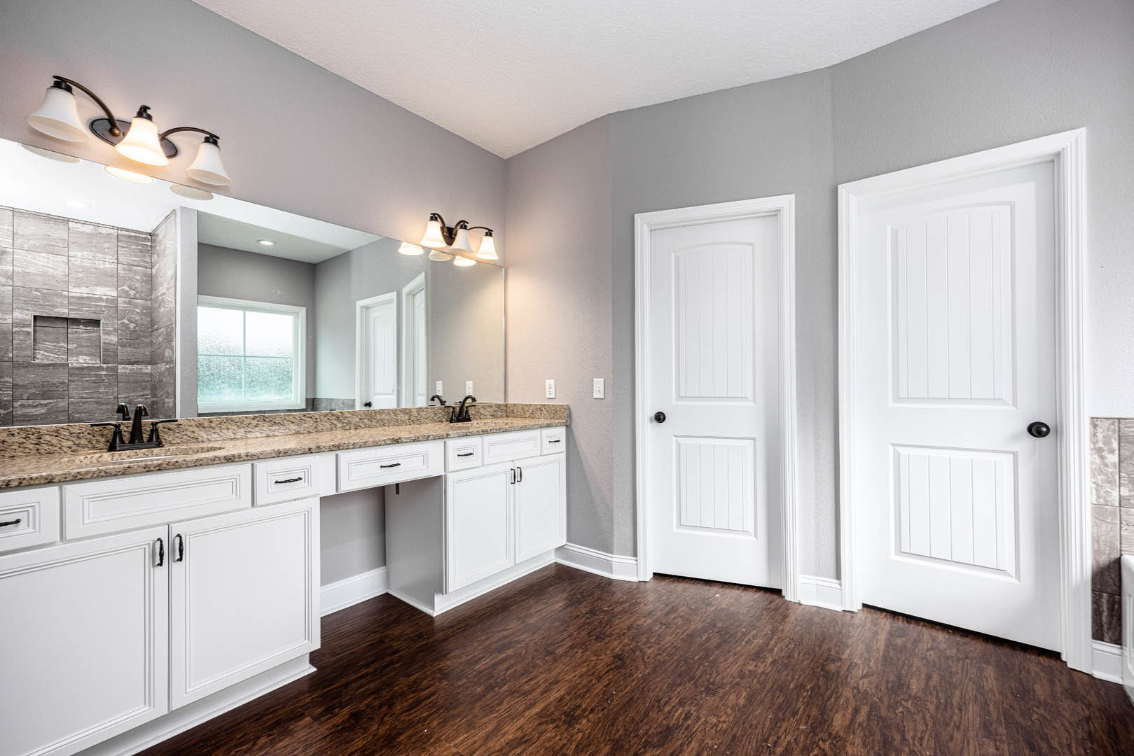 Bathroom with white shaker cabinets, quartz countertop, framed mirror above sink, frosted glass window, dark wood flooring with white baseboards, white door featuring black handle