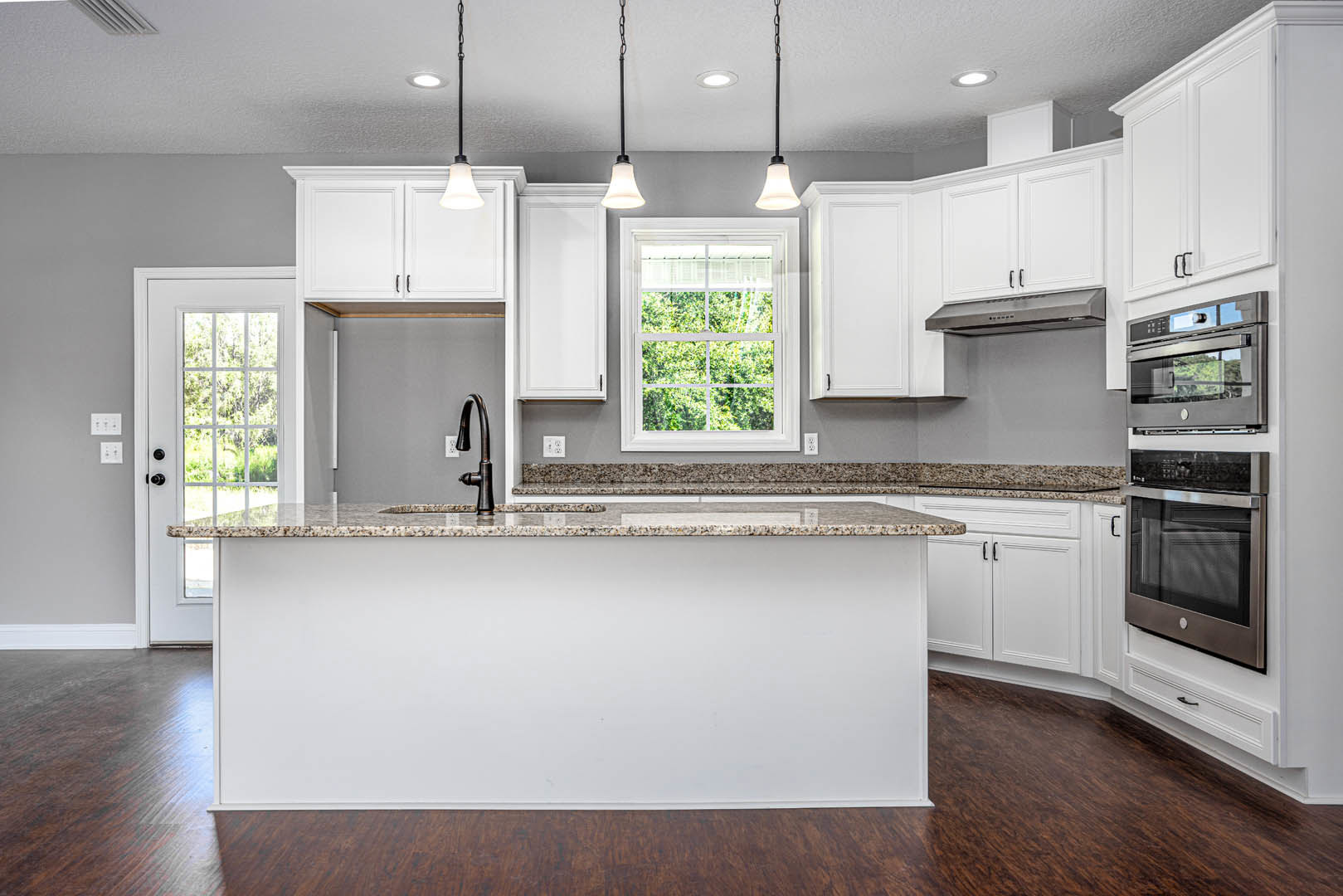 Marble kitchen countertop with white cabinetry, stainless steel microwave, chrome faucet, pendant light fixture, and window overlooking trees.