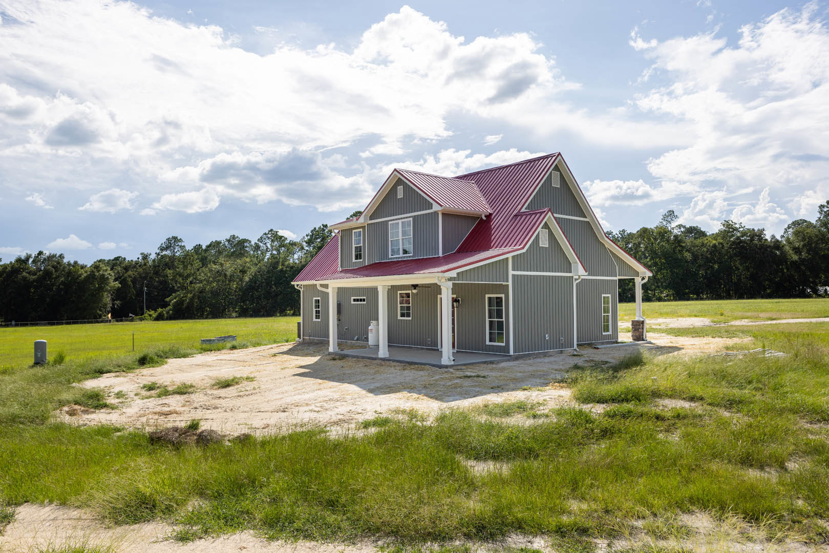 Red metal roof, white-framed windows, light-colored siding, surrounded by green grass and scattered trees under a partly cloudy sky