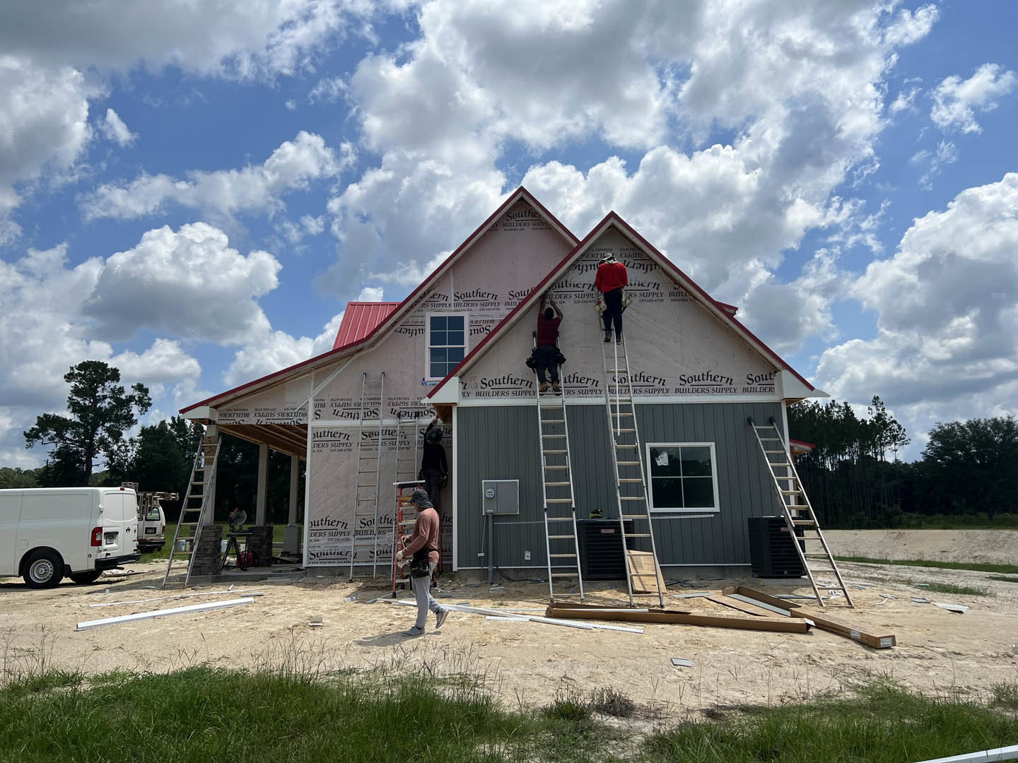 Workers on ladders installing or repairing siding on a two-story house with white van parked nearby, grassy yard, trees, and cloudy sky; window with ladder, person in red shirt