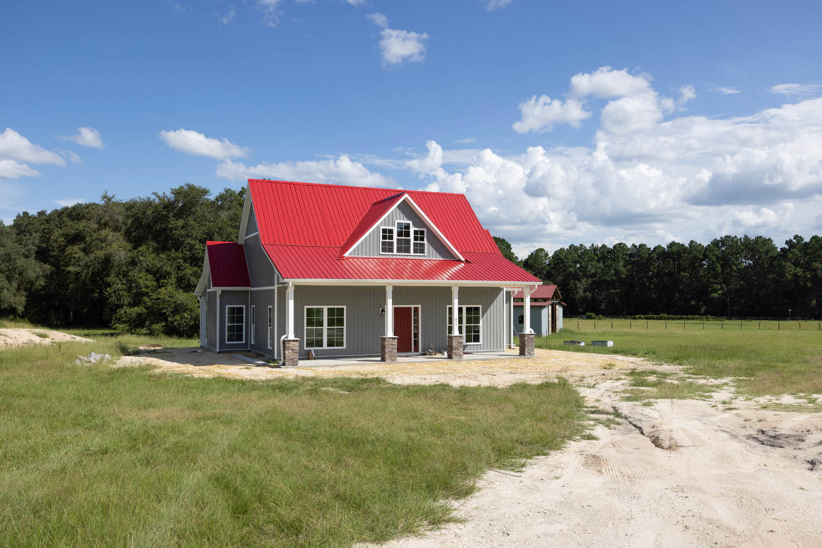 Red metal roof atop a white cottage-style home with white-framed windows, red front door, surrounded by green grass and mature trees under a blue sky.