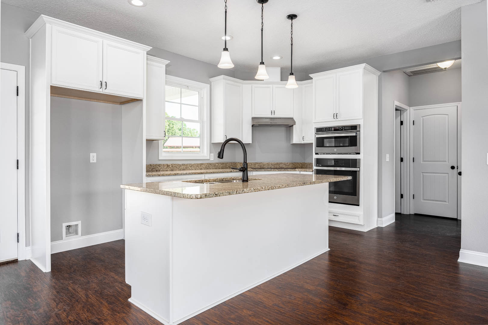 Marble kitchen island with white cabinetry, wood flooring, black faucet, built-in microwave and oven, white door with black hardware, pendant lights hanging from ceiling