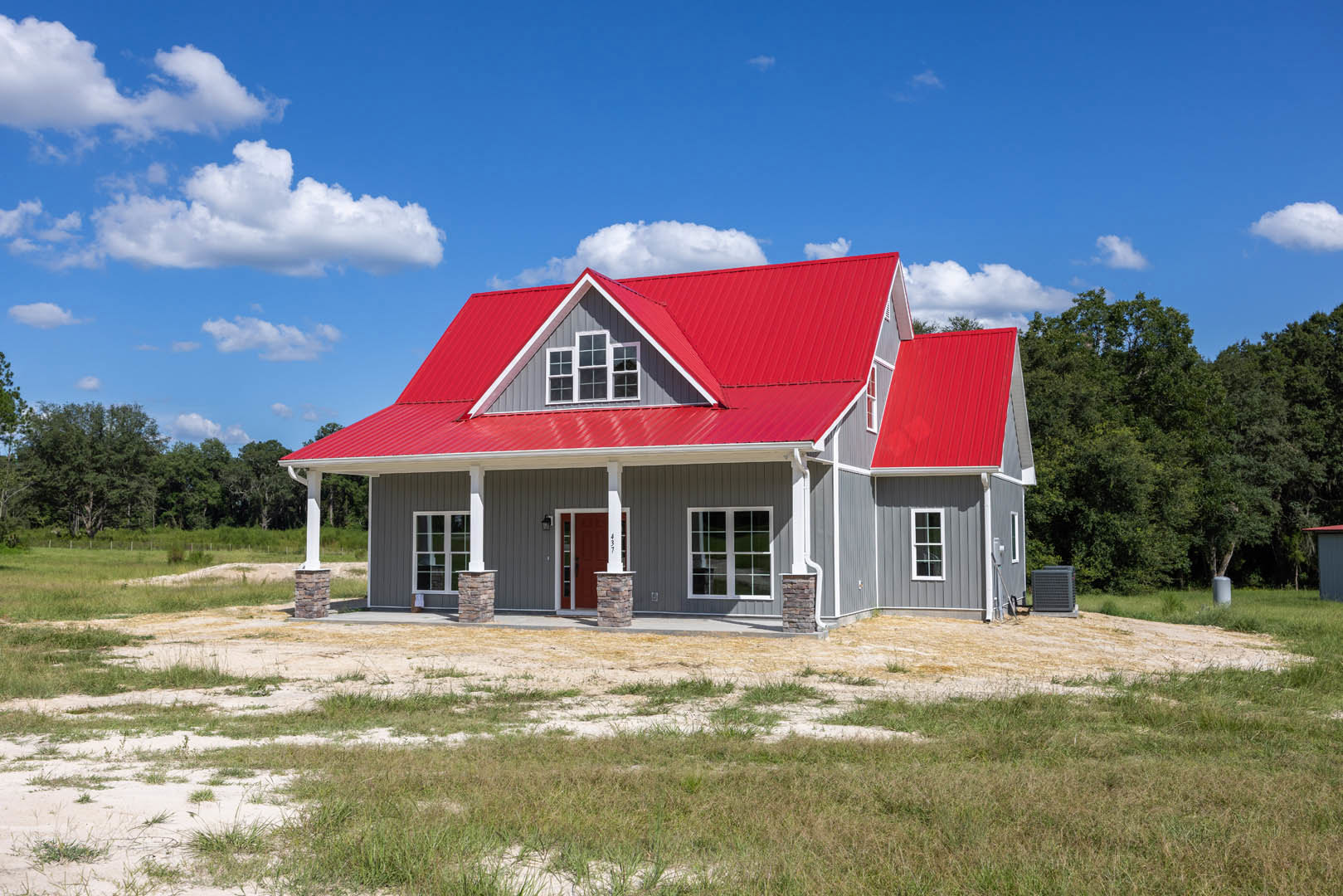Red metal roof atop a brick house with white-framed windows, grassy lawn in front, scattered trees, and partly cloudy sky overhead