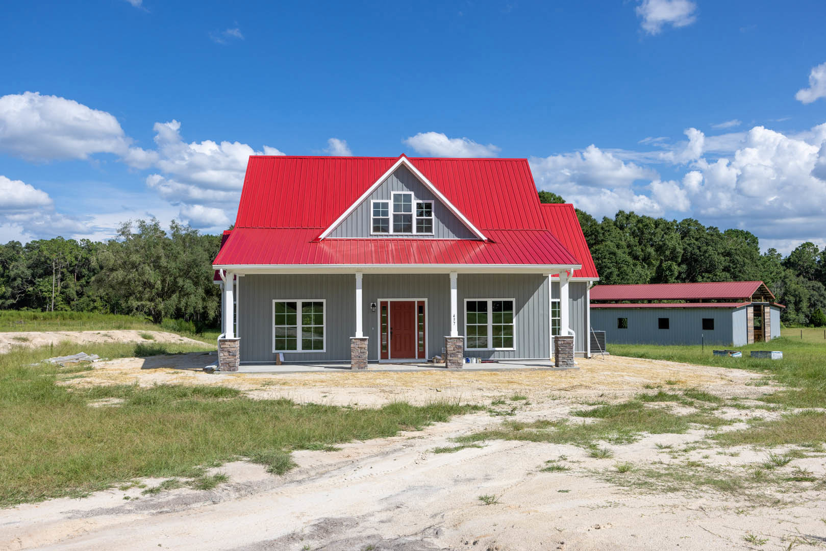 Red metal roof and multi-pane windows on a cottage-style home with a red glass-paneled door, surrounded by grass and trees, dirt driveway in foreground, cloudy sky overhead.