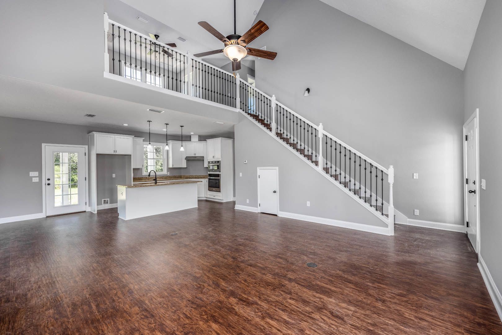 Open-concept living area featuring wood flooring, a central staircase with metal railings, white walls, a ceiling fan with light fixture, a kitchen with a sink, and a white door