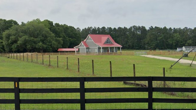 Red metal roof atop a white siding house, surrounded by green grass lawn, split rail fence, and leafy trees under blue sky; person standing near the front yard