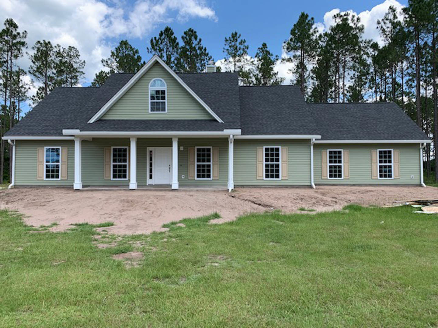 Two-story house with green roof, white siding, black-trimmed windows, white door with black square accents, front porch, lawn bordered by trees, cloudy sky overhead