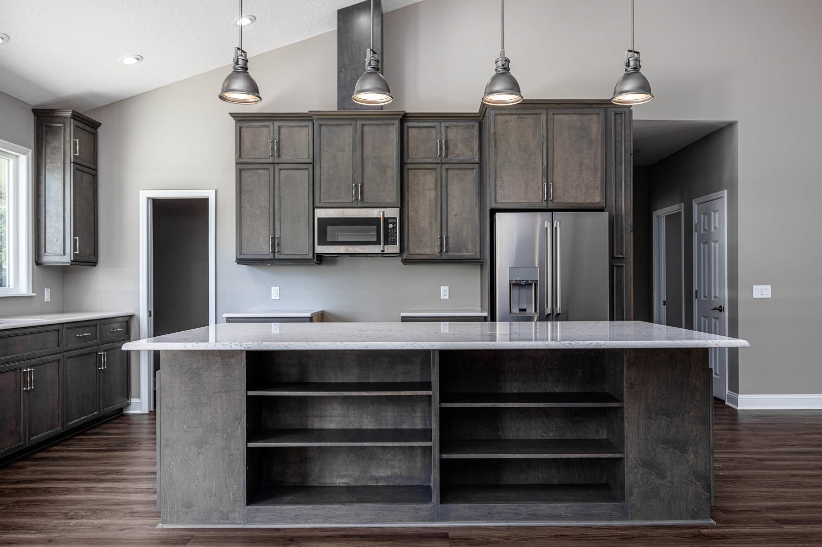 Spacious kitchen featuring a large marble-topped island with open shelving, white cabinetry, stainless steel refrigerator, farmhouse sink, and a window above the counter.