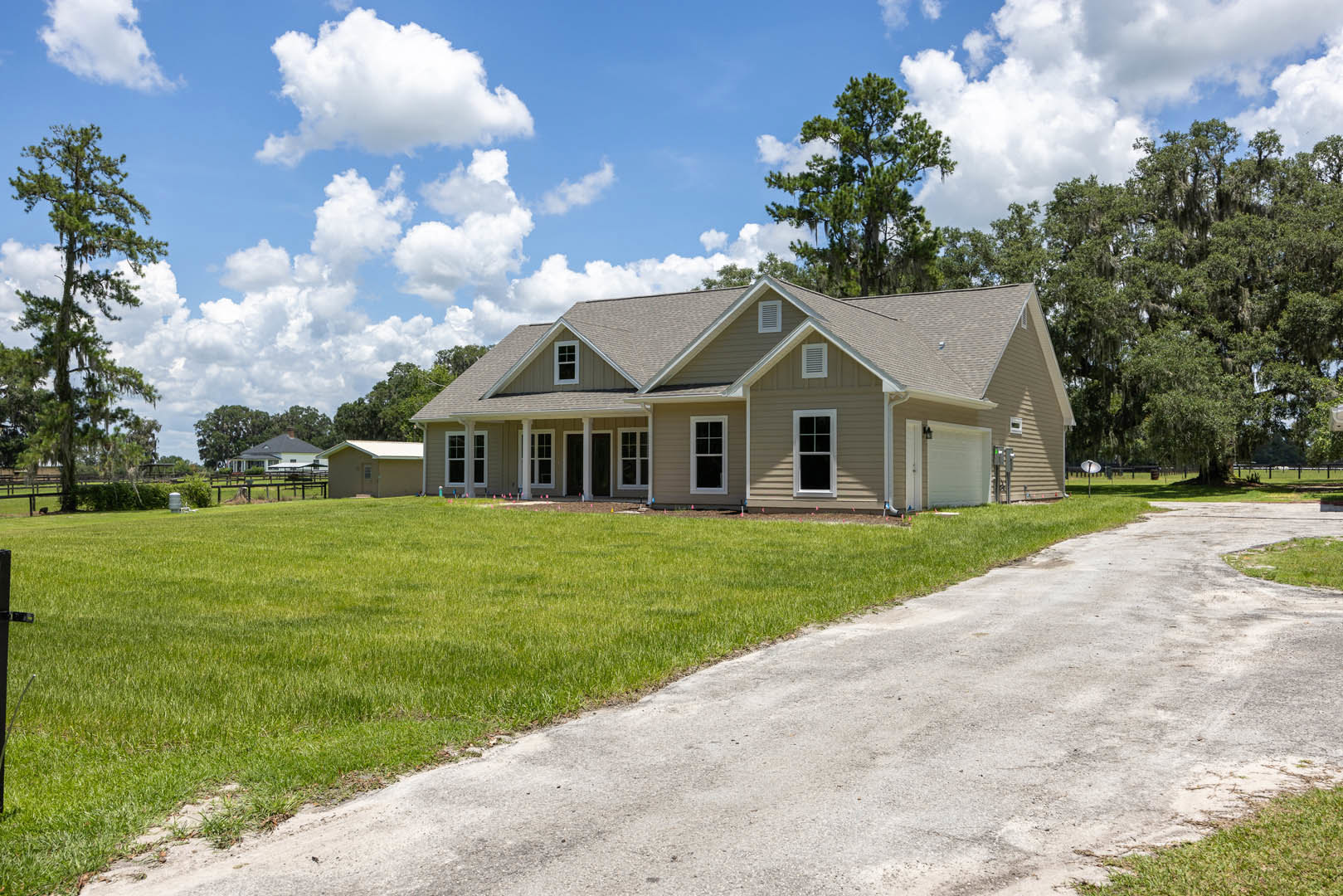 Two-story house with white siding, black-framed windows, paved driveway, manicured lawn, tall tree, and blue sky with scattered clouds