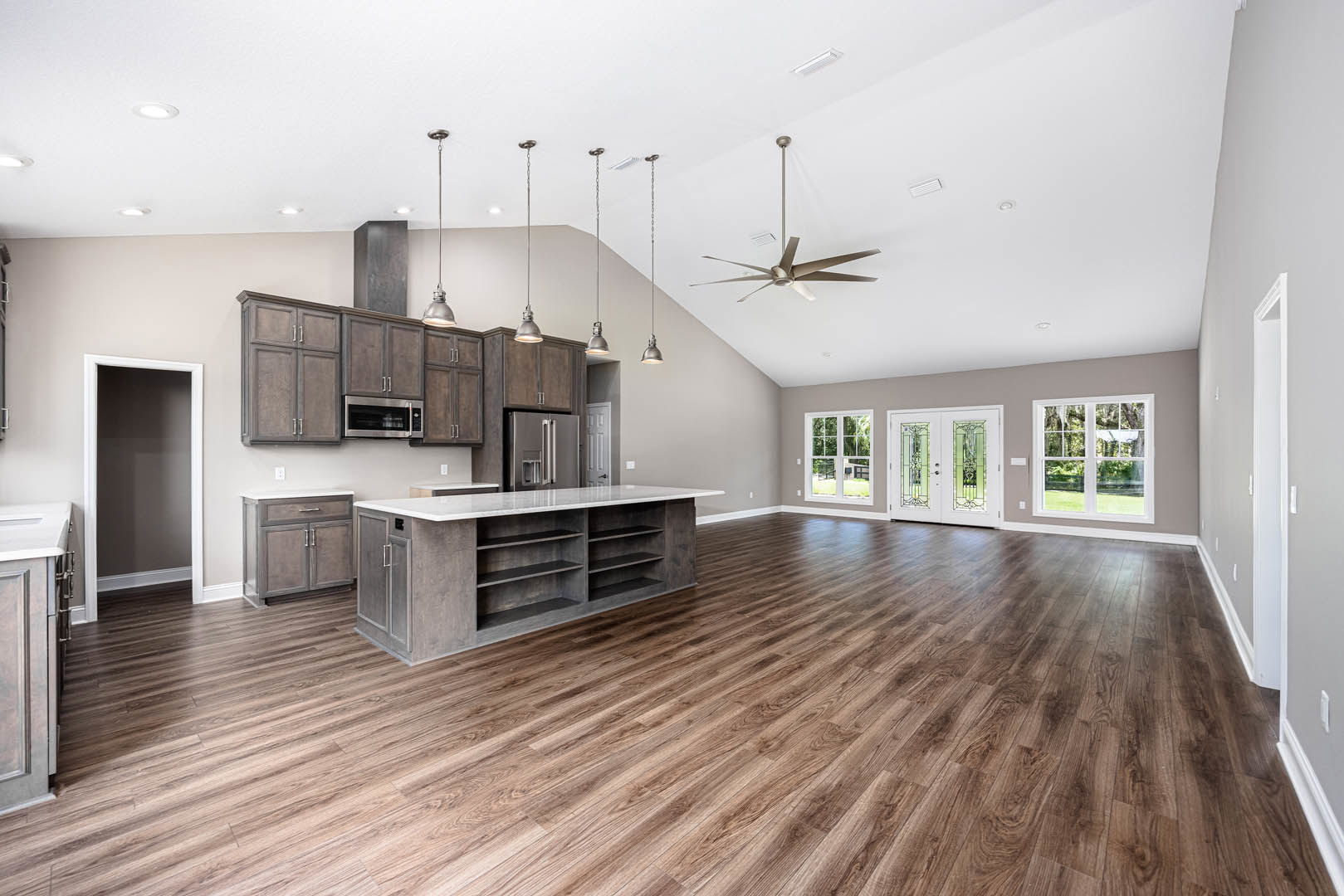 Spacious open floor plan featuring wood flooring, ceiling fan, kitchen counter with shelves, large window offering outdoor view, and white double doors with glass panels