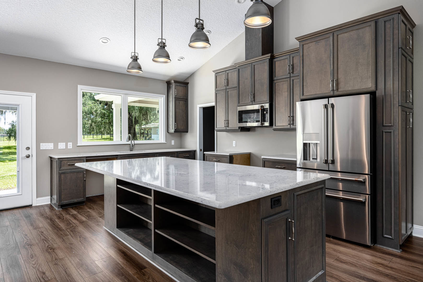 Spacious kitchen featuring a large marble island with open shelving, stainless steel refrigerator, white cabinetry, ceiling pendant lights, and a window framing a tree view.