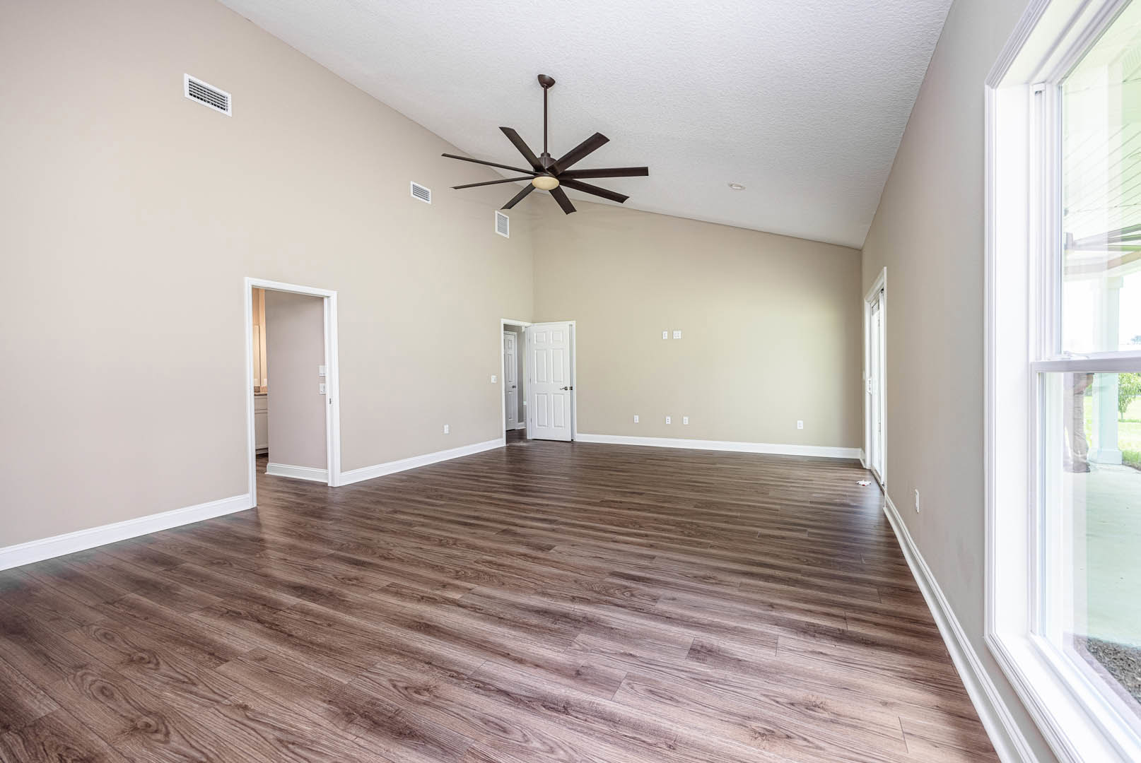 Spacious room with hardwood flooring, ceiling fan with light fixture, white door featuring silver handle, wall vent, and large window with white trim