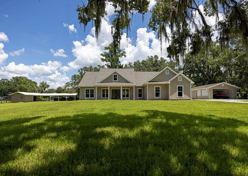 Two-story house with gray siding, covered front porch, attached garage, manicured green lawn, mature trees, and blue sky with scattered clouds