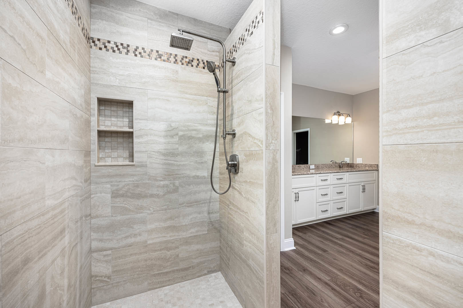 Bathroom with wood floor and cabinets, tile wall behind white shelf, ceiling light, window with dark background, metal shower head and rod.