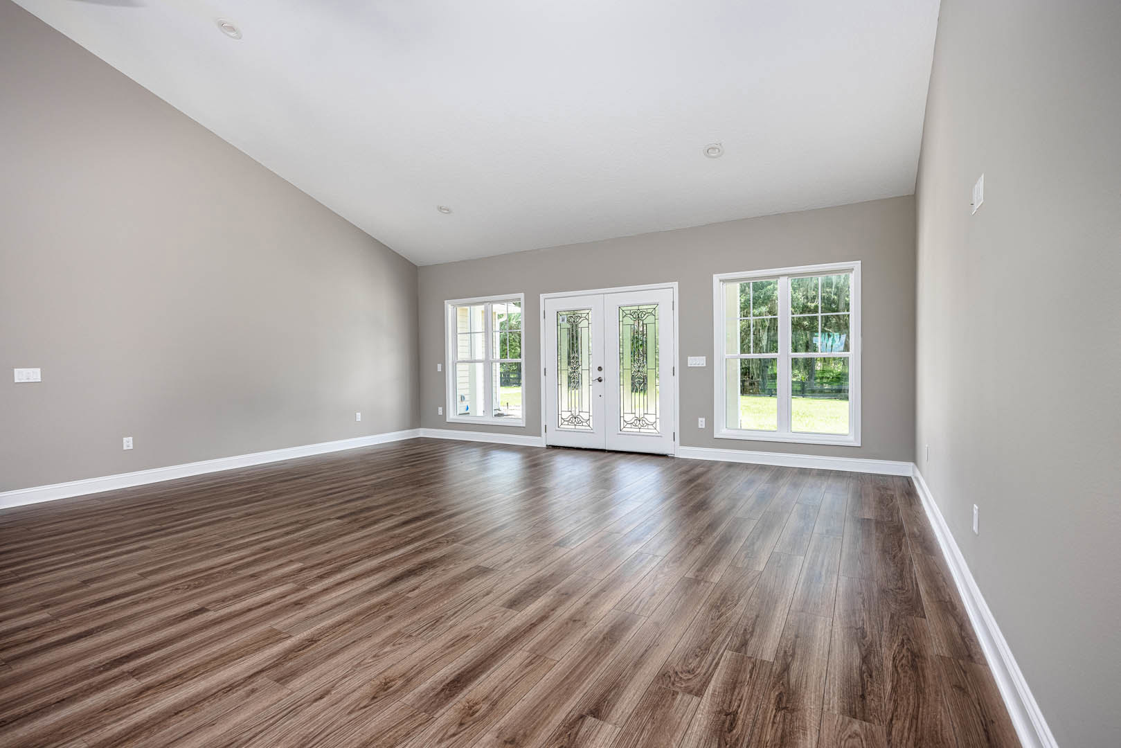 Hardwood floor in a bright room with white double doors featuring glass panels, large windows overlooking green lawn, smooth plaster walls, and ceiling.