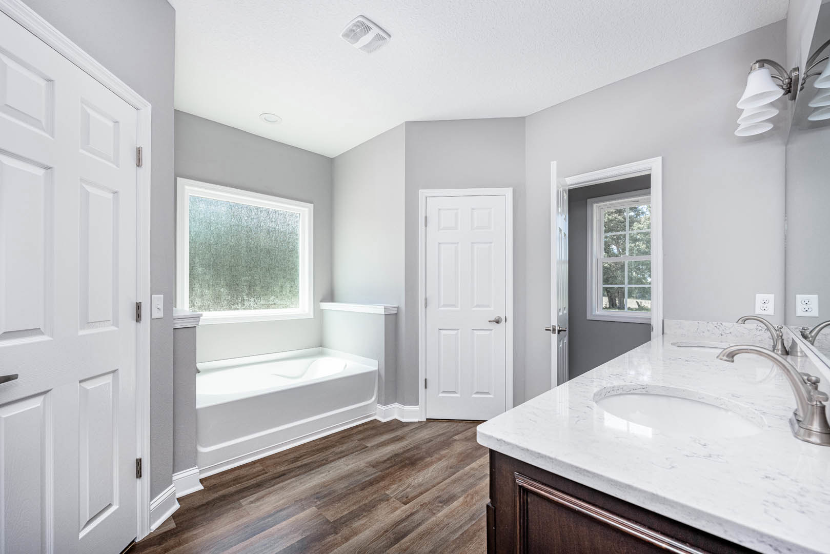 Bathroom with freestanding white bathtub, wood plank flooring, white countertop and sink, white door with silver handle, window above tub, neutral walls, modern fixtures