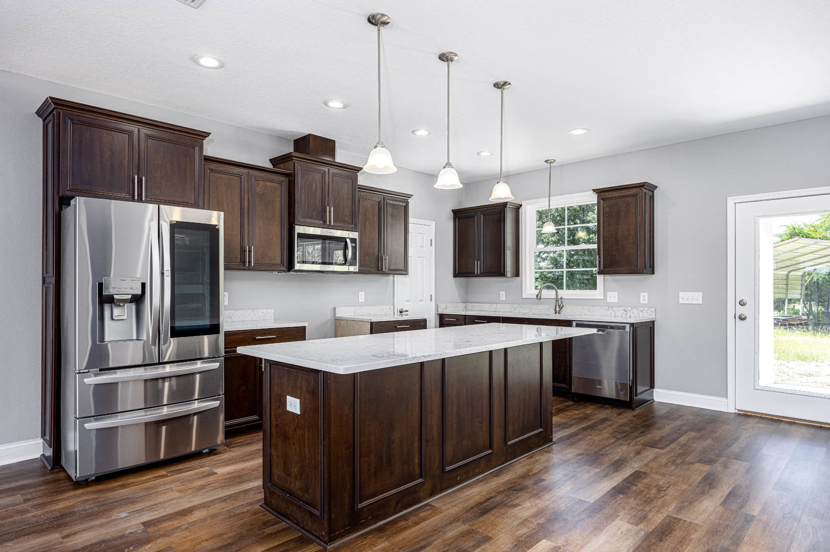 Dark wood kitchen cabinets, stainless steel refrigerator with glass door, marble-topped island, white door with metal handle, large window, light-colored flooring