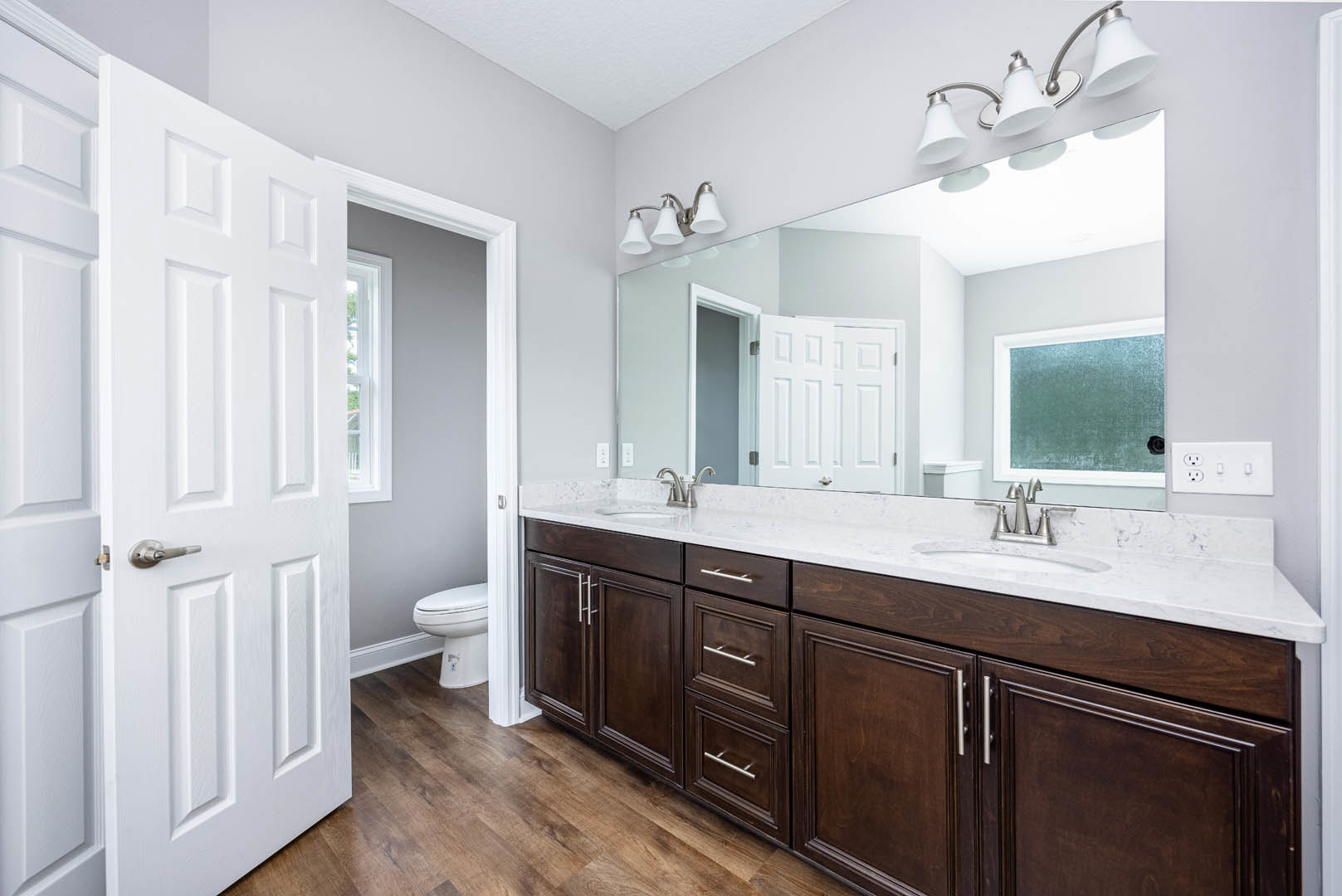 Bathroom with double sink vanity, large mirror above, brown cabinetry, chrome faucets, white toilet, tile flooring, window with natural light, modern light fixture