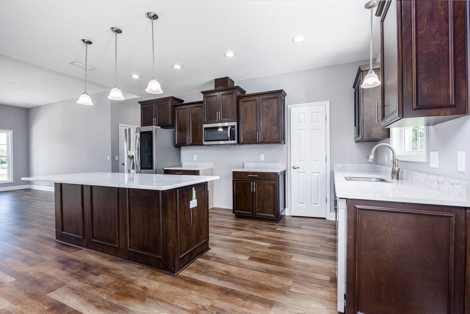 Kitchen with dark wood cabinetry, white marble countertops, central island, stainless steel appliances, white door with silver handle, wood panel accents, light fixture on ceiling