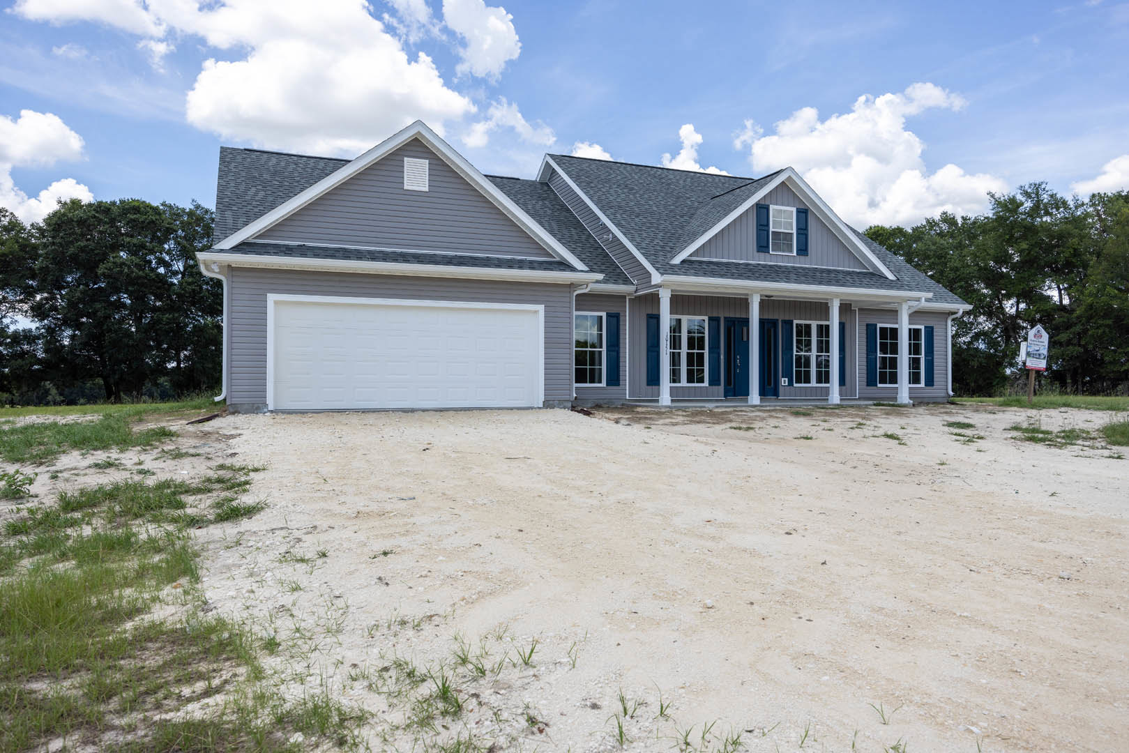 Two-story house with white garage door, gray shingle roof, dirt driveway bordered by grass, mature trees in background, cloudy sky overhead
