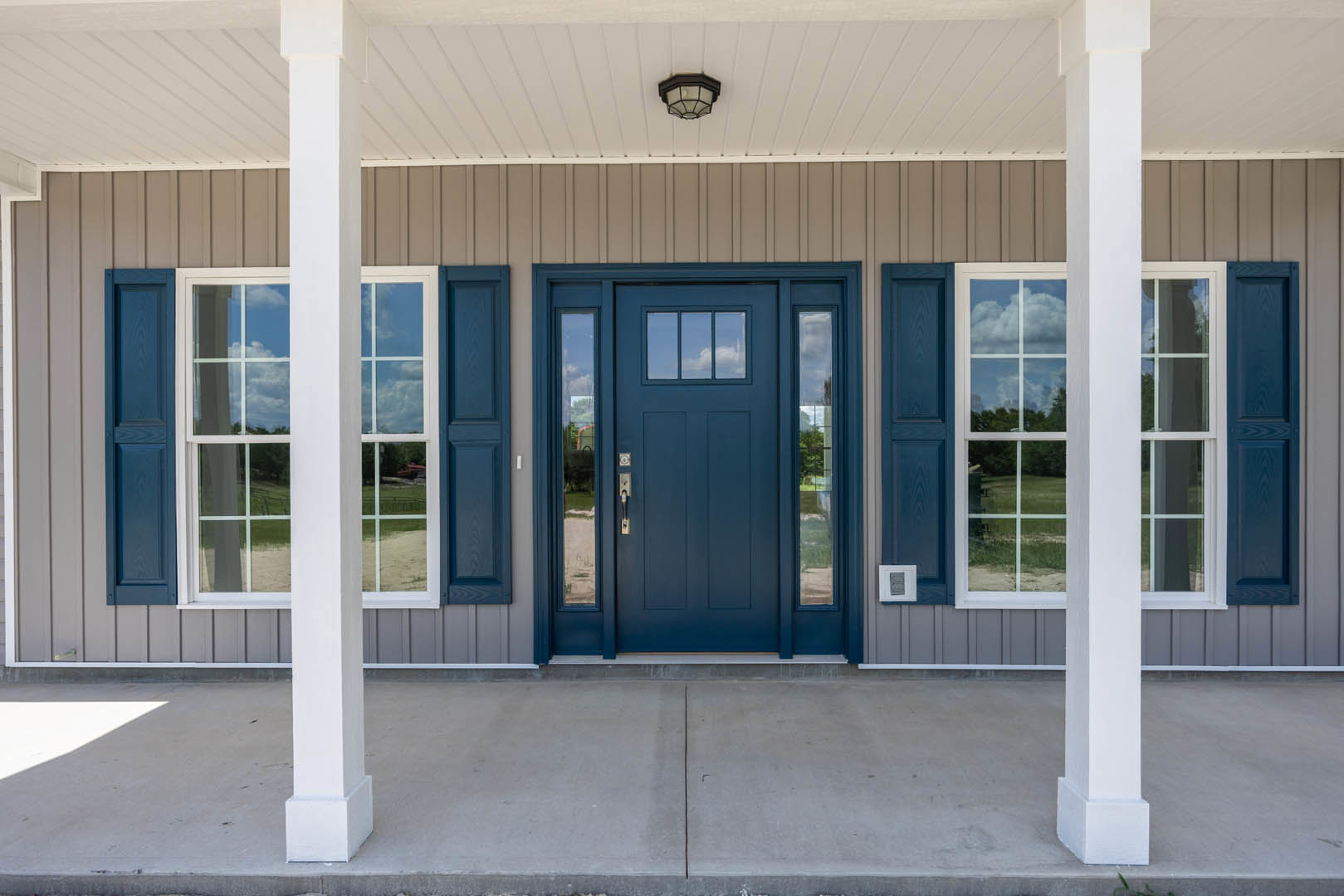 Blue front door with glass window, flanked by white square pillars, concrete entry floor, and ceiling light fixture; adjacent windows visible on house exterior.