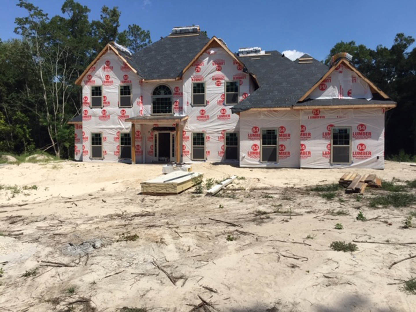 Partially built house wrapped in red and white protective sheeting, exposed wooden framing, white-framed windows, dirt yard with scattered construction materials and sticks